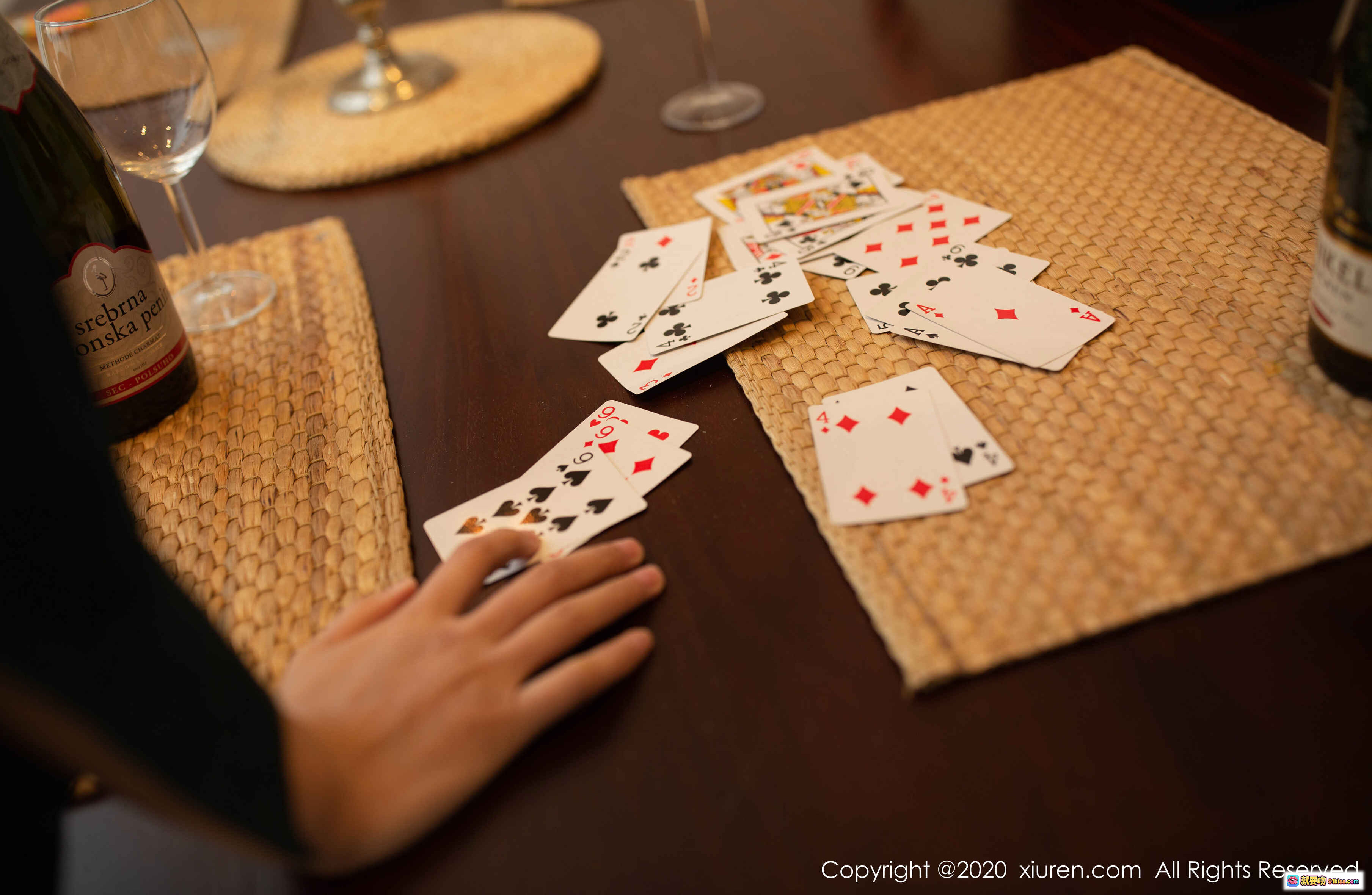 图片[1] - 桌游聚会场景：扑克牌散落木质桌面，手部特写搭配红酒酒瓶与高脚杯，编织餐垫营造温馨氛围，秀人网2020摄影风格 - 就要吻